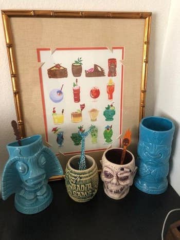 Four decorative tiki mugs are displayed successful  beforehand   of a bamboo framed poster featuring assorted  colorful cocktail illustrations