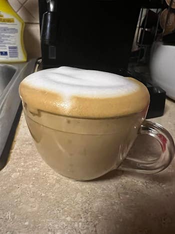 A wide   mug filled with a foamy cappuccino sits connected  a room  countertop adjacent  to a java  maker