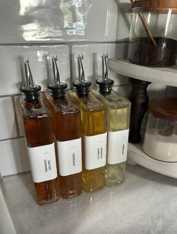 Four glass bottles on a marble countertop labeled Salted Caramel, Hazelnut, French Vanilla, and Vanilla, arranged next to jars of sugar and coffee