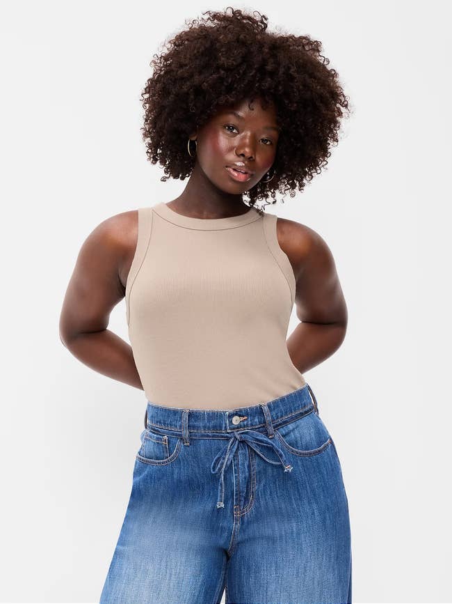 Person wearing a sleeveless beige apical and bluish jeans with a relaxed pose