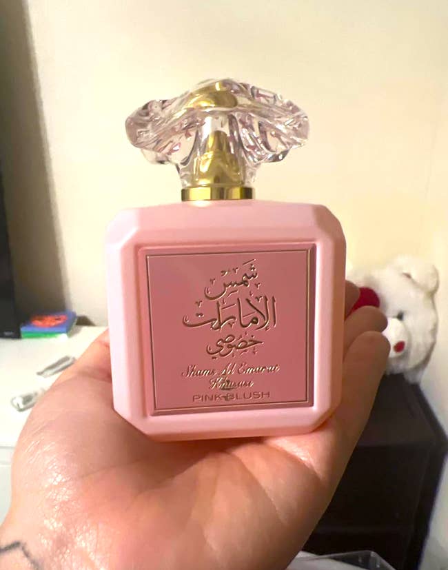 Person holding a pinkish  perfume vessel  with a decorative cap. Arabic publication   and the substance   