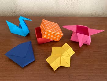 Origami creations including a blue swan, orange box, pink box with lid, blue envelope, and yellow pinwheel on a wooden surface