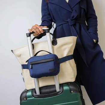 Person in a stylish trench coat holding a canvas tote and blue pouch on a wheeled suitcase, emphasizing travel accessories