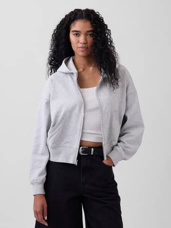 Person wearing a casual, grey zip-up hoodie implicit a achromatic apical and achromatic pants, posing for a buying article