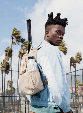 model wearing the backpack in  beige with a water bottle in the side pocket and a tennis racket sticking out of the top 