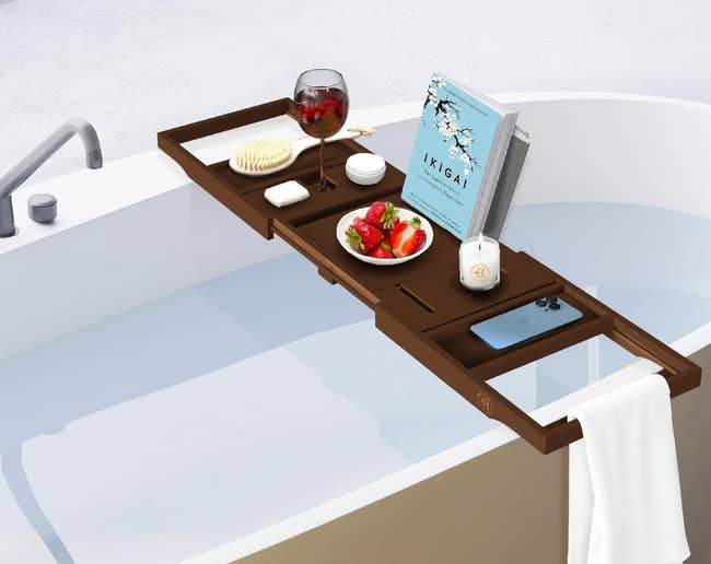 Expandable bathtub tray holding wine, a book, fruit, and bath items, extends from 27.75 to 41.5 inches