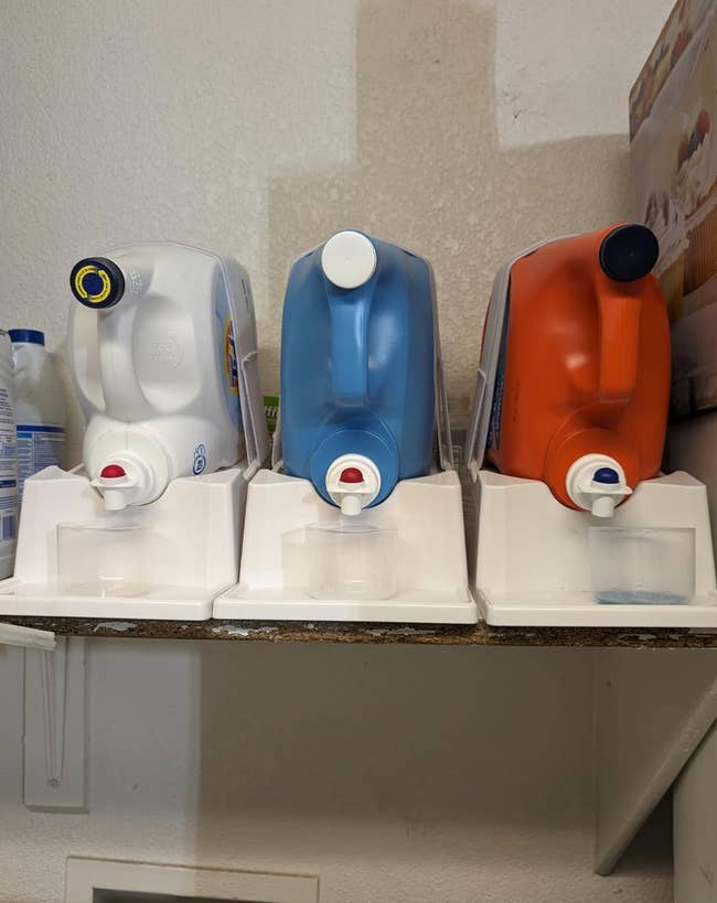 Three large detergent dispensers are on a shelf, each with a spout and cup underneath for dispensing liquid