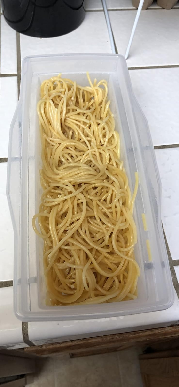 A clear container filled with cooked spaghetti noodles on a tiled kitchen counter