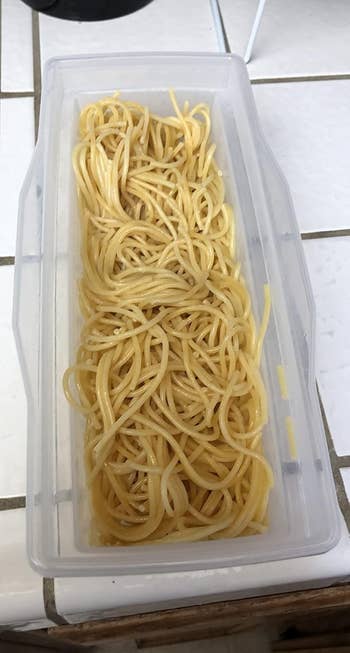 A clear container filled with cooked spaghetti noodles on a tiled kitchen counter