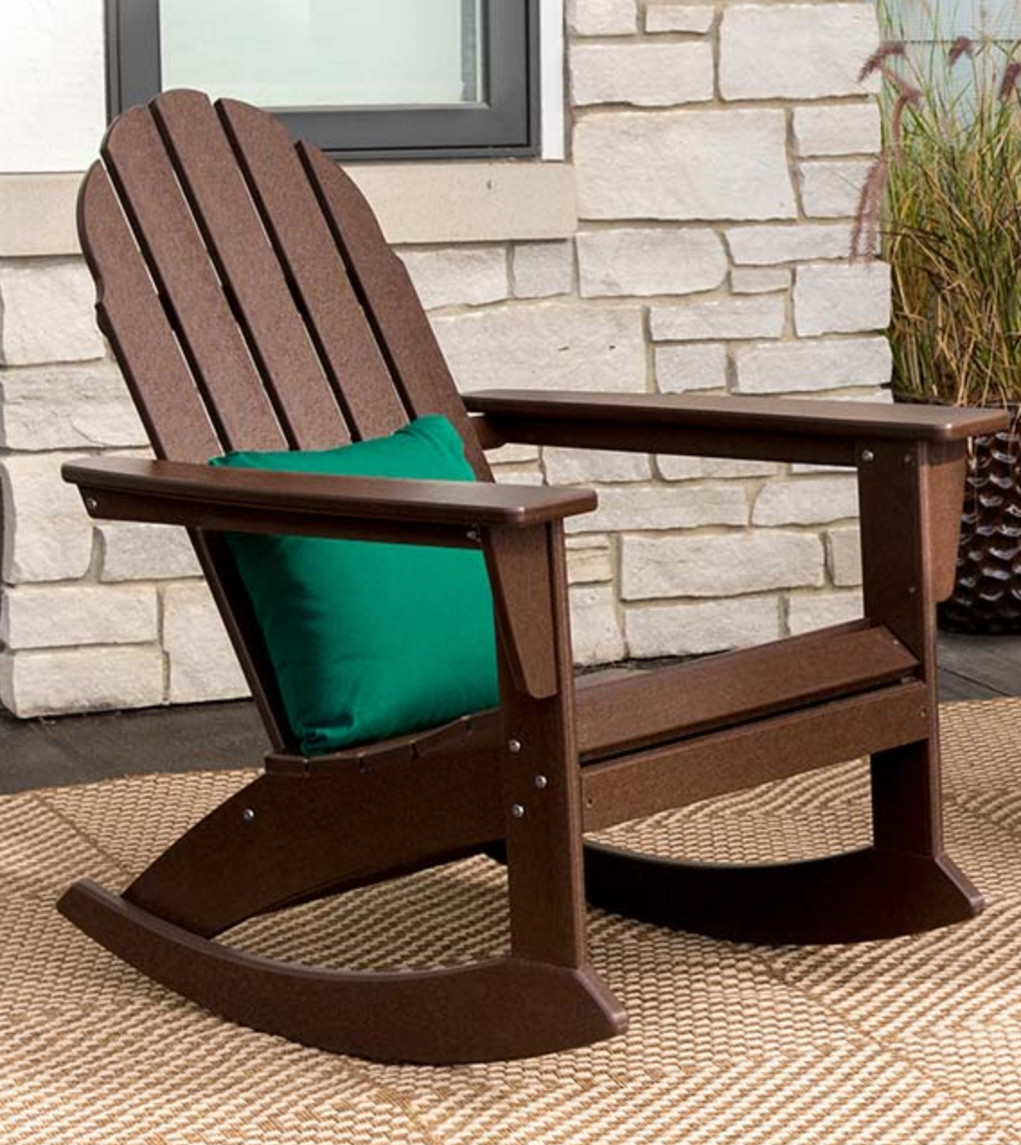 Dark brown wooden wide slatted rocking chair with curved back and straight arm rests, decorated with a dark green throw pillow outside on top of a striped rug