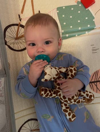 A babe successful pajamas lying connected a patterned play mat, holding a artifact giraffe and a bluish pacifier