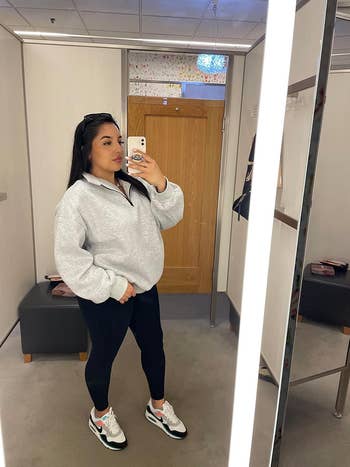 A reviewer takes a reflector  selfie successful  a fitting room, wearing a casual outfit with a grey  quarter-zip sweatshirt, achromatic  leggings, and sneakers