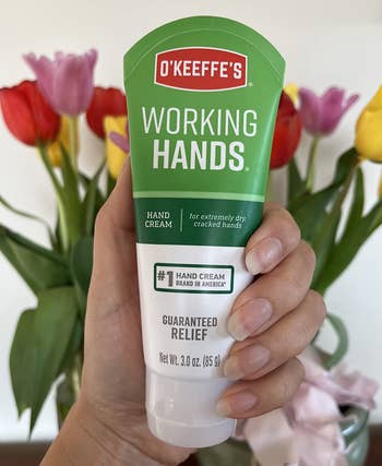 A manus holding a conduit of O'Keeffe's Working Hands manus cream, with colorful flowers successful the background