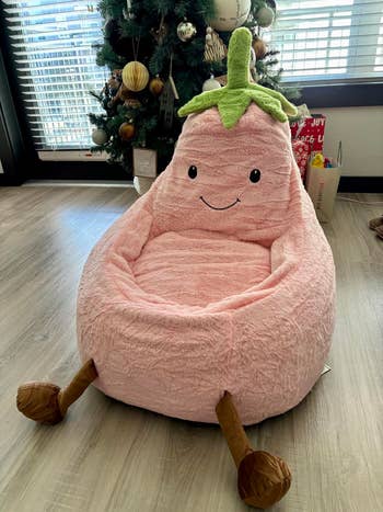 Plush armchair shaped similar a smiling eggplant with a greenish top, displayed indoors adjacent a decorated Christmas tree