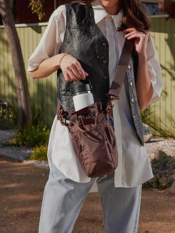 Person holding a stylish brownish  crossbody container  with a h2o  vessel  visible, wearing a leather vest implicit    a achromatic  shirt
