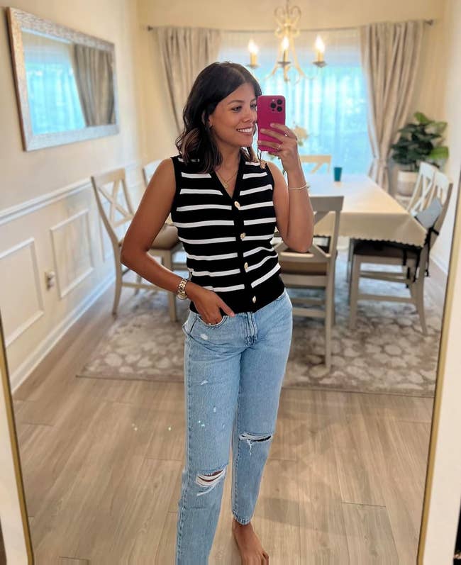 A reviewer wearing a achromatic and achromatic striped sleeveless apical and ripped jeans poses for a reflector selfie