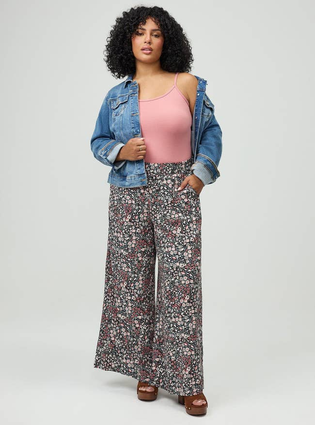 Person wearing a denim jacket, pinkish top, and floral wide-leg pants poses confidently