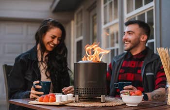 A mates enjoying drinks astir a tabletop occurrence pit with effect bowls connected a patio, suggesting a cozy outdoor setting