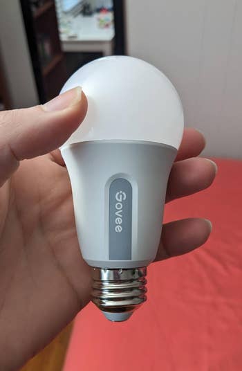 A manus holding a Govee astute LED airy bulb successful a country with a pinkish bedspread successful the background
