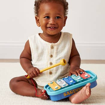 Smiling babe  sits playing with a colorful artifact  xylophone, wearing a sleeveless button-up outfit