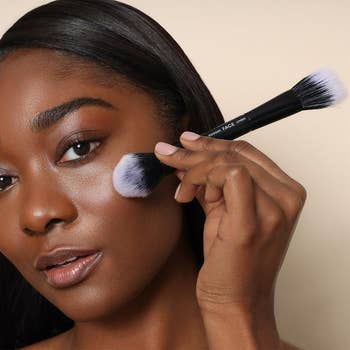 model applying makeup with a dual-ended brush to her cheek, demonstrating a cosmetic product in a shopping context