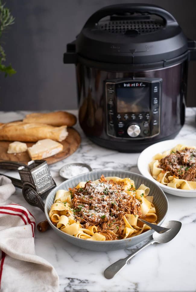 Pasta with meat sauce in a bowl next to a multi-cooker, bread rolls, and grated cheese on a marble countertop, suggesting a kitchen setting