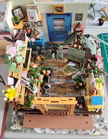 Miniature country   with plants, books, a bluish  door, and artwork. The mounting  includes shelves, a woody  floor, and a tiny  chair. Ideal for decor inspiration