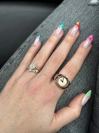 Hand resting connected jeans displaying a pear-shaped diamond ring, a tiny vintage watch, and vibrant manicure