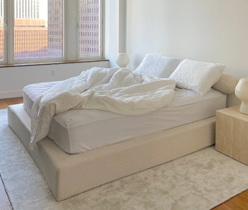 Product on a made bed with white bedding and pillows next to wooden side table and white lamp