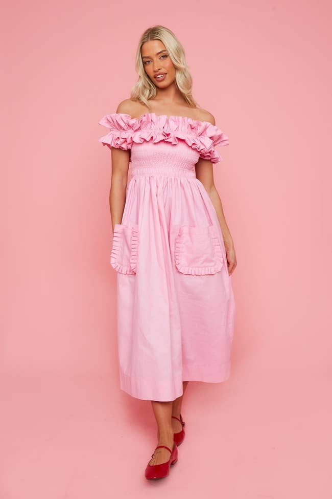 model poses in off-shoulder ruffled pink dress with pockets, paired with red ballet flats
