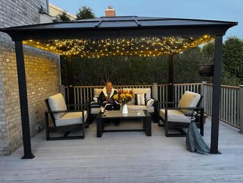 Person sitting nether  a platform  gazebo decorated with drawstring  lights, surrounded by cushioned seating and a array  with a colorful bouquet