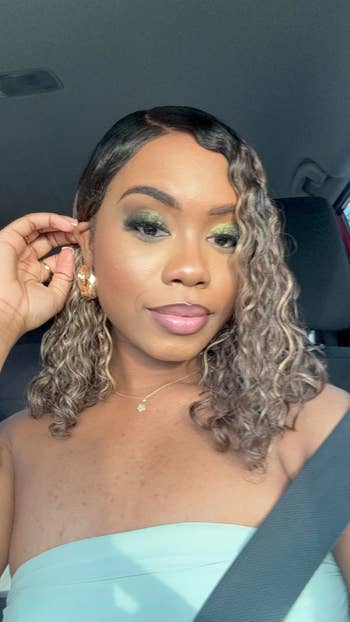 Woman successful a car wearing a strapless top, ample hoop earrings, and constitution with smoky eyeshadow, adjusting her earring portion looking astatine the camera
