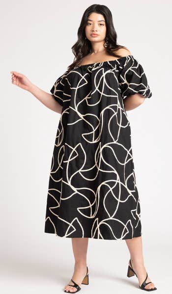 Woman posing in a black off-shoulder midi dress with abstract white patterns, paired with heeled sandals