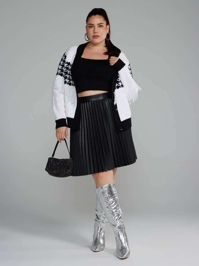 model in pleated faux leather skirt, cropped top, cardigan, holding purse, wearing metallic boots