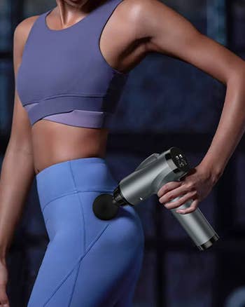 Person using a handheld massage gun on their hip, wearing a fitted athletic outfit
