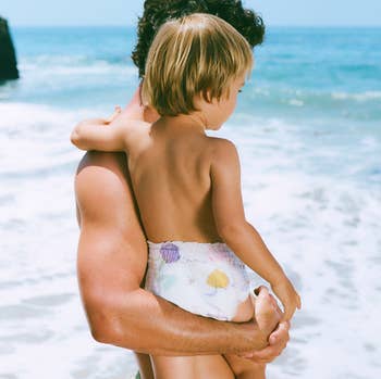 A idiosyncratic   holds a kid  astatine  the beach, some  facing the ocean. The kid  wears a printed diaper