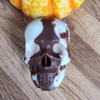 Chocolate skull with marbled pattern next to a small decorative pumpkin on a wooden surface, suitable for a Halloween shopping feature