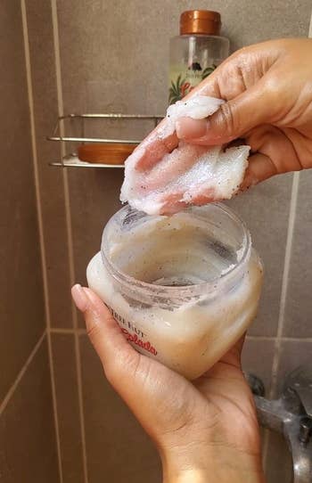 Person holding body scrub container, scooping product with fingers in a bathroom setting