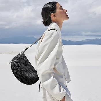 A fashion model poses outdoors in a minimalist white outfit with a black textured handbag over her shoulder, showcasing a stylish look for a shopping article