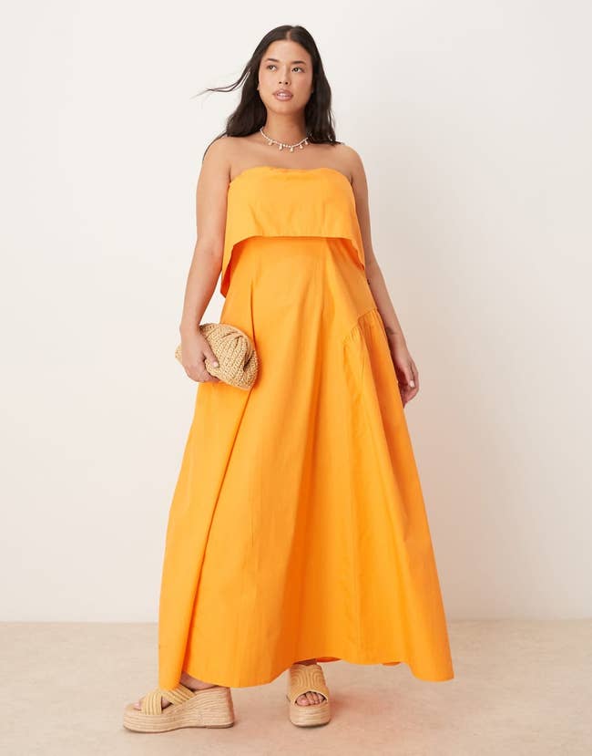 A model poses in a flowy strapless dress with a layered top, holding a woven clutch, wearing platform sandals, highlighting a breezy, summer style