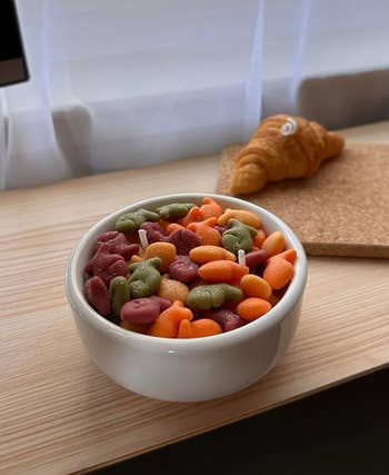 A white bowl filled with colorful fish-shaped crackers on a wooden surface with a croissant in the background