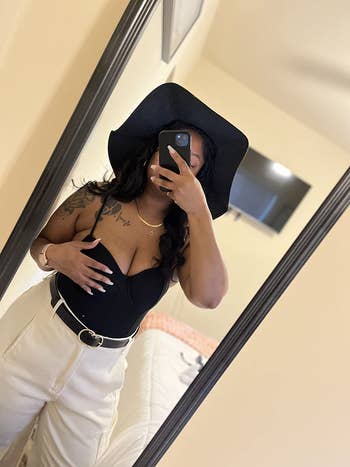 Person wearing a strapless achromatic top, wide-brimmed hat, and high-waisted pants takes a reflector selfie