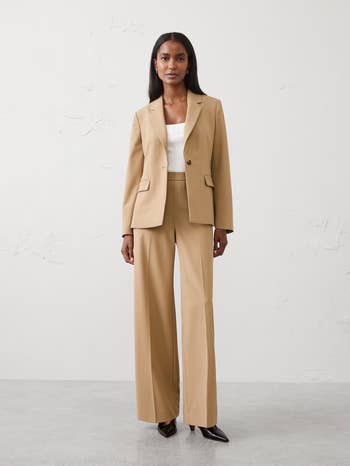 Model wearing a tailored tan suit   with a achromatic  apical  and achromatic  heels, lasting  against a neutral backdrop