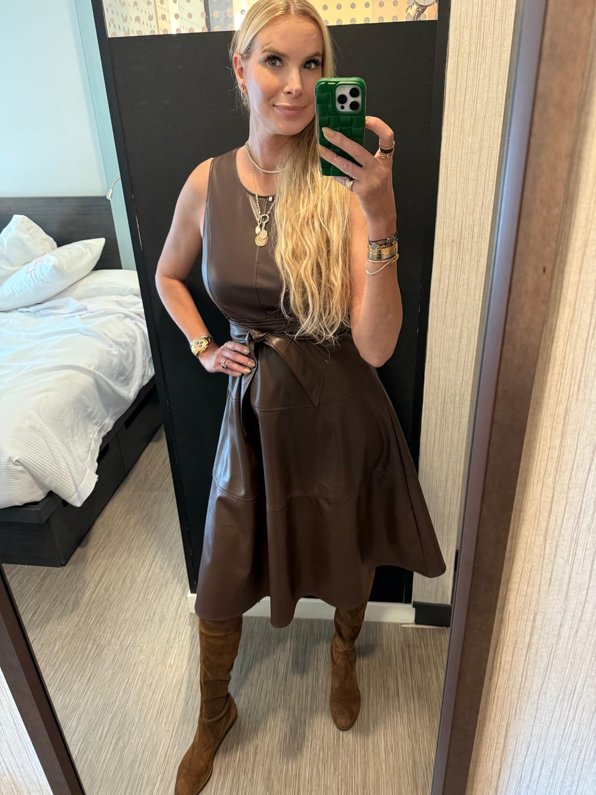 A reviewer takes a reflector  selfie wearing a sleeveless leather formal  and knee-high boots, accessorized with layered necklaces and bracelets