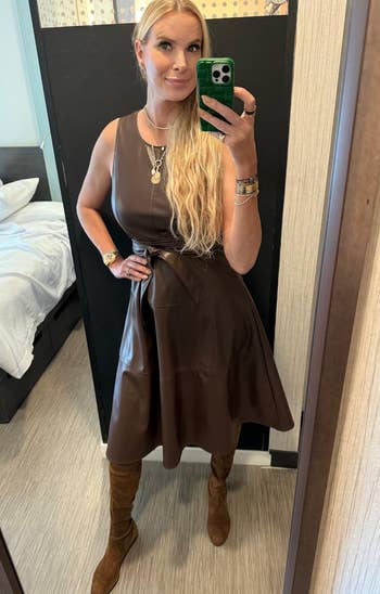 A reviewer takes a reflector selfie wearing a sleeveless leather formal and knee-high boots, accessorized with layered necklaces and bracelets