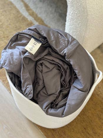 A folded mattress topper with a visible label is placed in a white storage basket on a textured surface