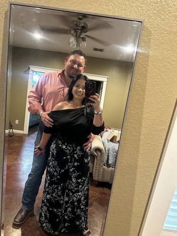 A couple takes a mirror selfie at home. The woman wears an off-shoulder top and floral pants. The man is in a button-up shirt and jeans