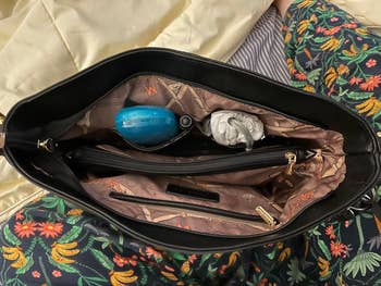 Open handbag with floral lining, containing a blue perfume bottle, a white object, and a zipped compartment. Set against a floral patterned fabric