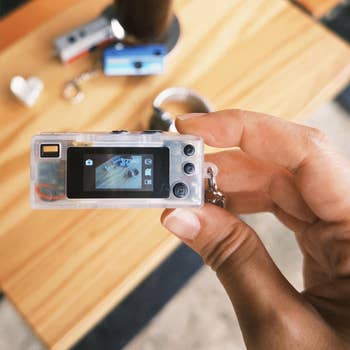 Person holding a small digital keychain camera with a wooden table and various items blurred in the background
