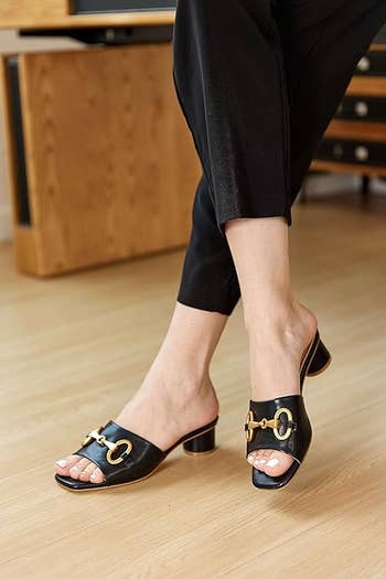 Person wearing black heeled sandals with gold chain detail, highlighting the shoe design for shopping context